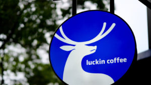 close up luckin coffee's (LKNCY) logo on light box hangging outside of coffee booth. Blur green trees background.
