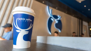 A Luckin (LKNCY stock) coffee cup on a table in a Luckin shop with the logo on the wall behind.