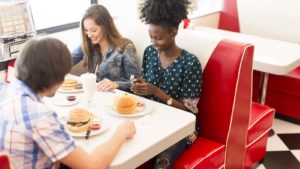 Three young adult friends sit around a vintage restaurant booth eating hamburgers. image represents restaurant stocks