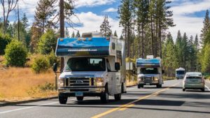 Two RVs drive down a road with trees and blue sky in the background.