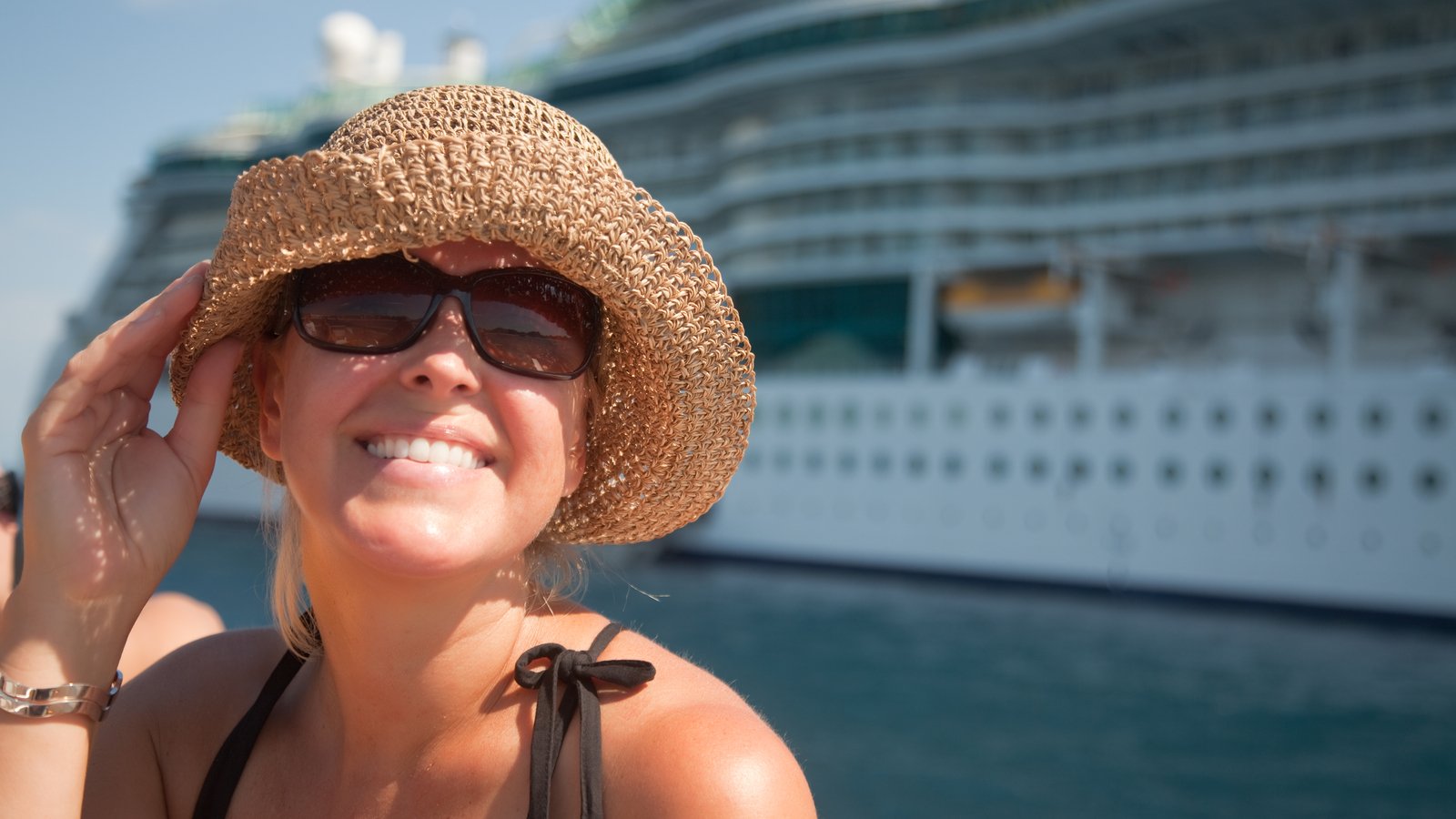 A close-up shot of a woman wearing a straw hat with a cruise ship in the background representing cruise stocks.