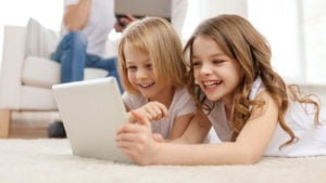 An image of two young girls looking at a tablet and smiling while an adult reads in the background. represents gnus stock