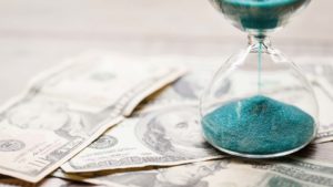 An hourglass filled with blue sand sits on top of $100 bills.
