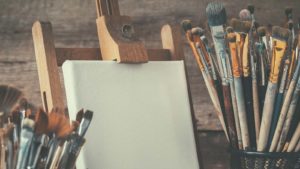 A wooden easel holds a white canvas and is surrounded by bins of different size paintbrushes. 