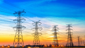 multiple powerline towers are shown against a sunset and a distant city skyline. AQN stock
