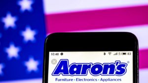 The logo for Aaron's (AAN) is displayed on a smartphone screen with an American flag in the background.