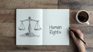 A close-up shot of a hand doodling "Human Rights" in a notebook next to a drawing of a balanced scale.