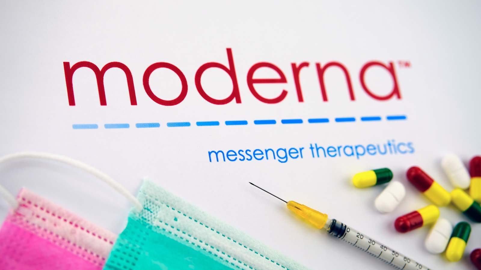 The Moderna (MRNA) logo surrounded by syringes, pills and disposable face masks.