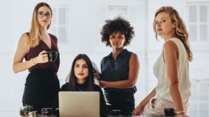 A group of four women in business attire pose around a desk.