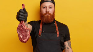 Look, its delicious. Self confident male butcher holds raw ribeye steak, suggests to buy for low price, has tattooed arm, long red beard, wears apron and gloves. Chef carries prime rib, going to bake.