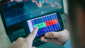 Stocks to buy: smartphone with the words "buy" and "sell" displayed on the screen. The user's finger is about to press buy. Stock charts are in the background of the image.