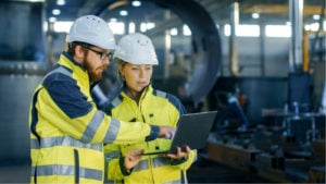 Engineers in yellow jackets and hard hats looking at a laptop in a workshop