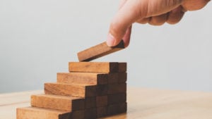 a hand laying another wooden block as the top step on a miniature staircase of wooden blocks