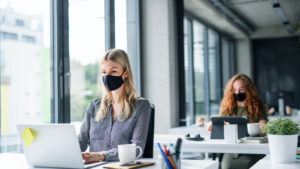 two women working on laptops in office space with black PPE face masks on