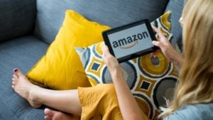 Woman sitting on couch using tablet with "Amazon" on display.