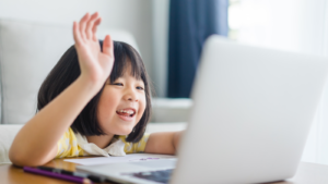 Image of a young girl raising her hand in front of a laptop.