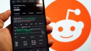 A smartphone shows GameStop (GME) up 70% with the Reddit logo in the background representing Meme Stocks Rally.