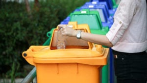 person depositing a plastic water bottle in a yellow plastic recycling bin. The trash can is in a line of several other blue and green trash cans.