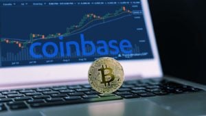 A Bitcoin rests on top of a computer with the Coinbase (COIN) logo and a trading chart. 