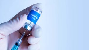 A gloved hand holds a syringe inserted into a bottle labeled 'Coronavirus Vaccine' on a light blue background.
