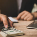 hands at desk near laptop computer, with one hand holding a pile of hundred dollar bills. Bank stocks