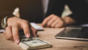 hands at desk near laptop computer, with one hand holding a pile of hundred dollar bills