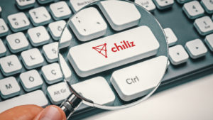 Hand holding a magnifying glass over a keyboard key that has the Chiliz altcoin logo on it