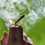 photo of a hand holding a marijuana joint that is smoking against a green outdoor background