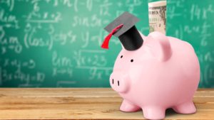 A pink piggy bank wearing a grad cap sits on a wooden table in front of a chalkboard.