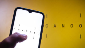 Canoo (GOEV stock) logo displayed on smartphone screen as well as in background on yellow wall