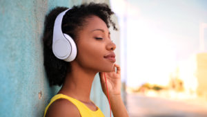A woman listens to music on headphones while standing against a wall in an outdoor environment.