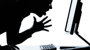 A shadowy figure looking at a computer screen has a look of rage on their face.