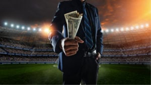 A person wearing a suit and tie holds a handful of dollar bills in the middle of a brightly lit sports stadium.