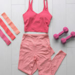 A pink athleisure outfit is laid out on a floor next to a pair of dumbbells.