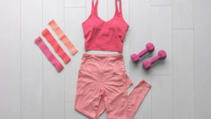 A pink athleisure outfit is laid out on a floor next to a pair of dumbbells.