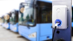 An electric vehicle charger is seen next to a row of blue electric buses. ARVL makes electric vans and buses.