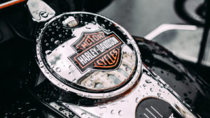 A close-up photograph of the tank of a Harley-Davidson motorcycle with raindrops on it.