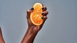 A hand with black nail polish squeezing an orange in front of a grey background.