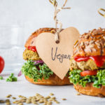Two plant-based burgers are placed next to a variety of vegetables. One of them has a heart-shaped label hanging off of it with the handwritten word "vegan" on it. vegan stocks