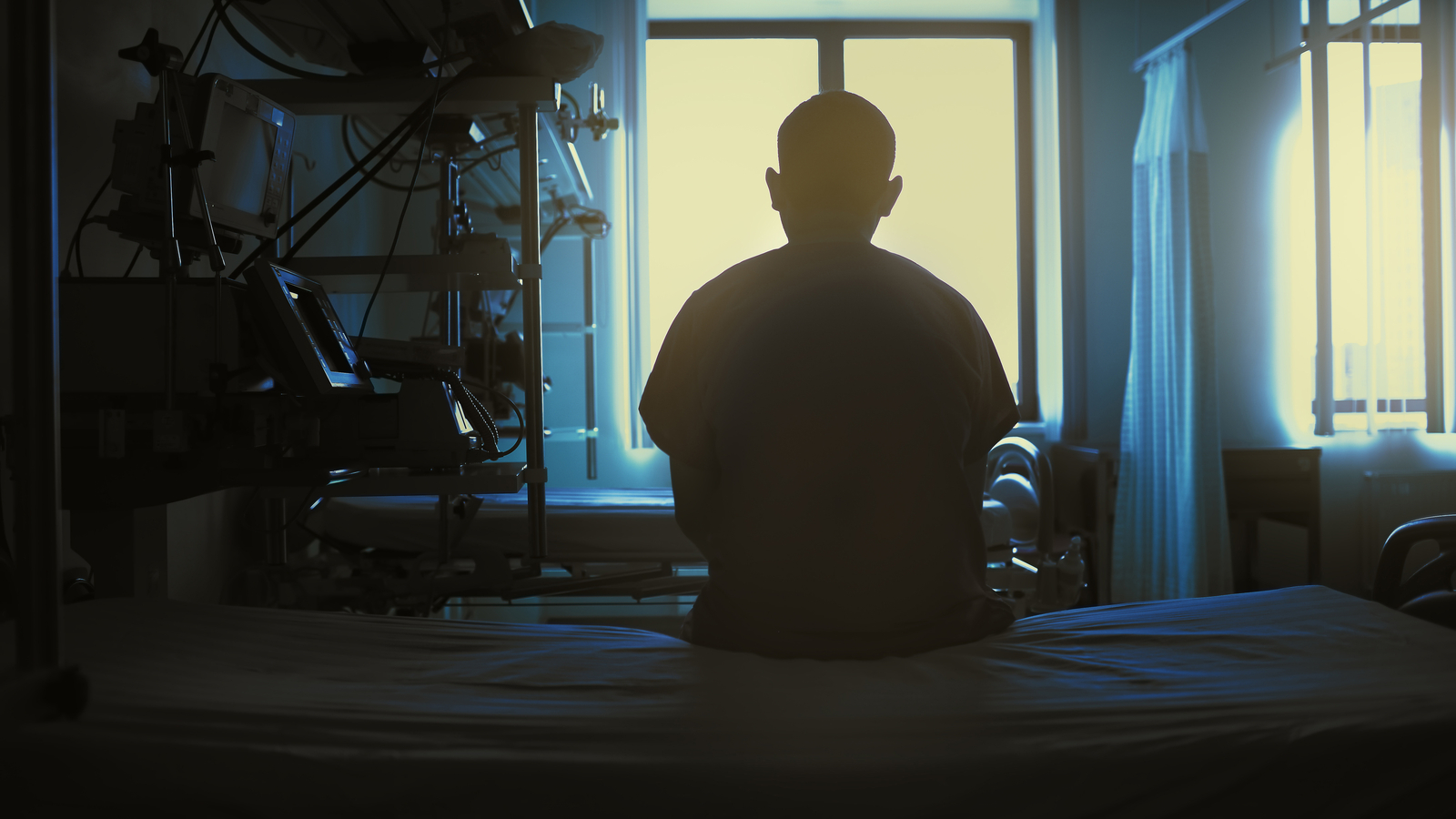 A person in silhouette faces toward a bright window while sitting at the edge of a hospital bed.