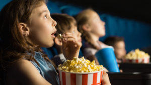 girl looking fascinated eating popcorn watching a movie at the local movie theatre representing movie stocks.