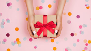 GiftCrowd: two hands holding a gift that is wrapped in brown paper and a red ribbon over a pink background with multicolored confetti