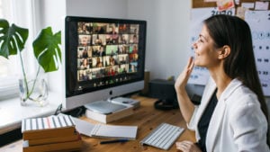 A woman sits at a desk and waves to a large number of people on the video conferencing software Zoom (ZM).