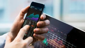 A person holds a phone with a stock chart visible on it with another chart visible on a computer nearby.