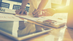 Two people take notes on paper in a brightly-lit room with a laptop, a tablet, and a phone all on the same table.