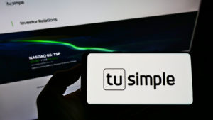 TSP stock: a hand holding a phone displaying the Tusimple logo in front of a computer displaying the company's investor relations page 