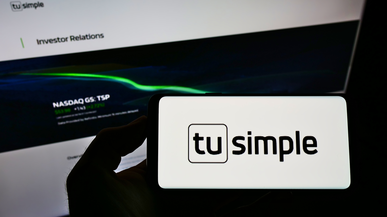 TSP stock: a hand holding a phone displaying the Tusimple logo in front of a computer displaying the company's investor relations page