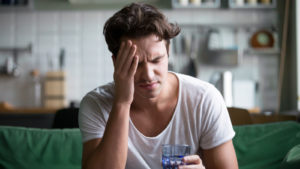 A tired-looking person holds a hand to their forehead and holds a glass of water.