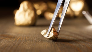 A photo of a gold nugget on a table taken with tweezers, with more gold behind it.
