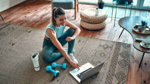 A woman uses a laptop computer on a mat with a pair of dumbbells and a water bottle on the floor beside her.